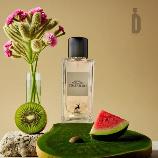 A bottle of Maison Al Hambra L'Impressio Iii 100ml EDP fragrance is displayed against a beige background. The elegant setup includes a green glass vase with fluffy green and pink flowers, a half-sliced kiwi, and a watermelon slice on a green, textured surface.