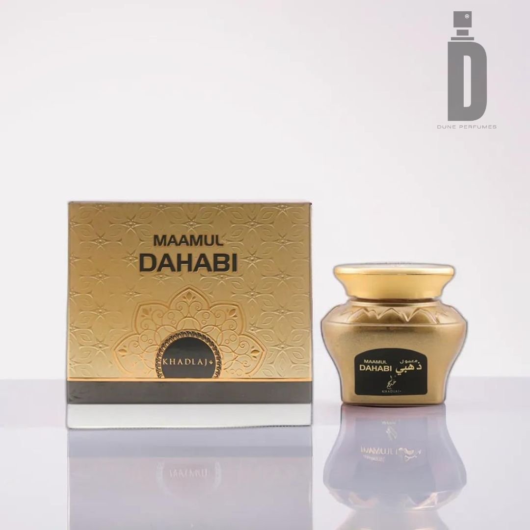 A gold-colored box labeled "Maamul Dahabi Bakhoor 48g - Khadlaj" with intricate designs sits next to a matching gold jar adorned with a black label, evoking a sense of luxury and tradition. The items rest on a reflective surface against a blurred white background, while the "Dune Perfumes" logo graces the upper right corner.