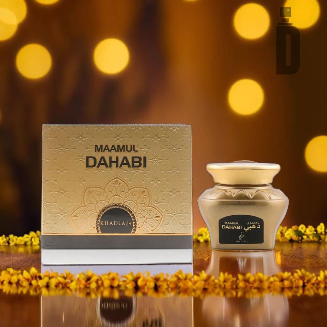 A product shot featuring a box and a jar labeled "Maamul Dahabi Bakhoor 48g" by Khadlaj. The gold box adorned with floral motifs and the elegantly shaped jar with a matching gold lid evoke luxury and tradition. The background is illuminated with warm, golden bokeh lights and small yellow flowers, highlighting the allure of perfumed oud wood chips.