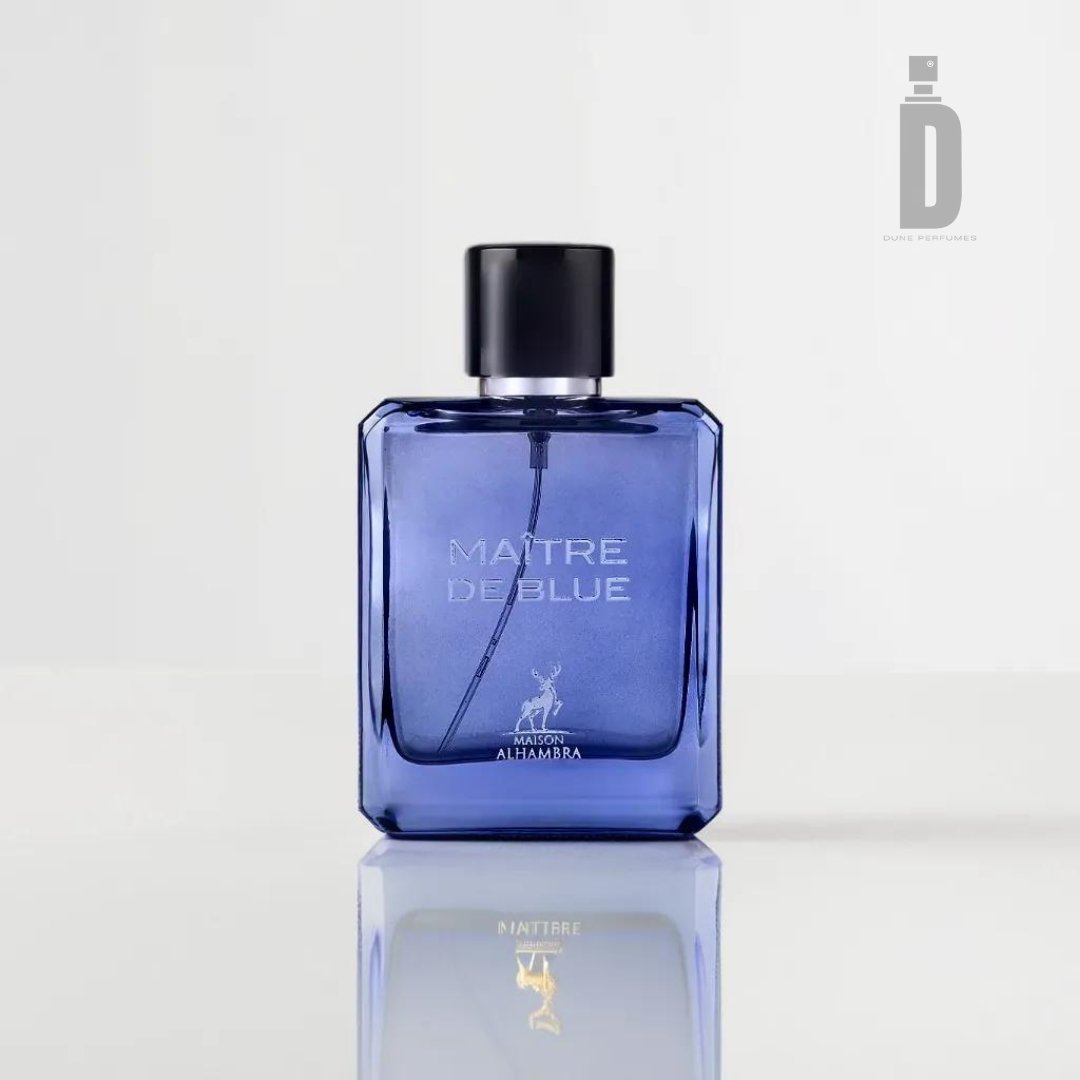 A blue rectangular perfume bottle with a black cap, labeled "MAÎTRE DE BLUE" on the front. The label also includes a stag logo and "MAISON ALHAMBRA" underneath. This captivating fragrance from Maison Al Hambra exudes timeless elegance, and the white background casts a faint reflection of the bottle at the base.