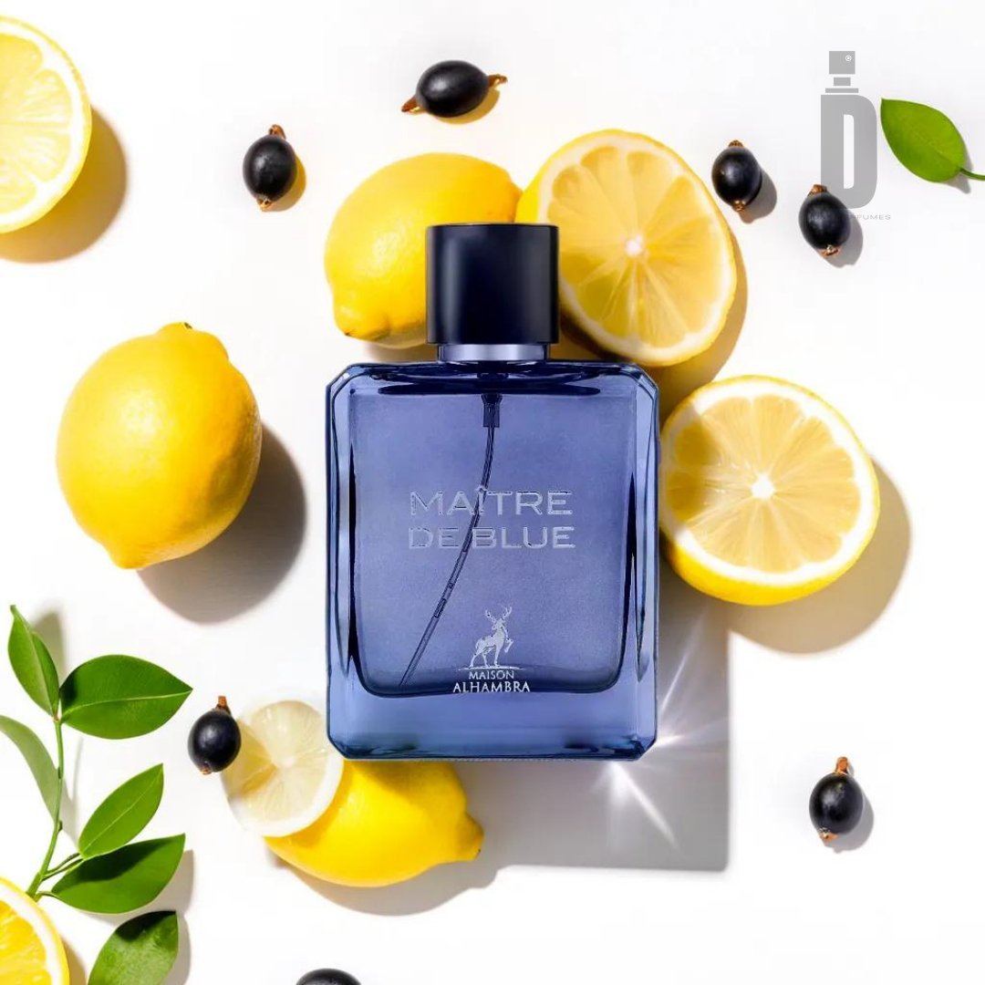 A blue perfume bottle labeled "Maitre De Blue 100ml EDP - Maison Alhambra" by Maison Al Hambra is surrounded by halved and whole lemons, black currants, and green leaves on a white background. The centrally positioned bottle, exuding timeless elegance, casts a faint shadow under the sunlight.