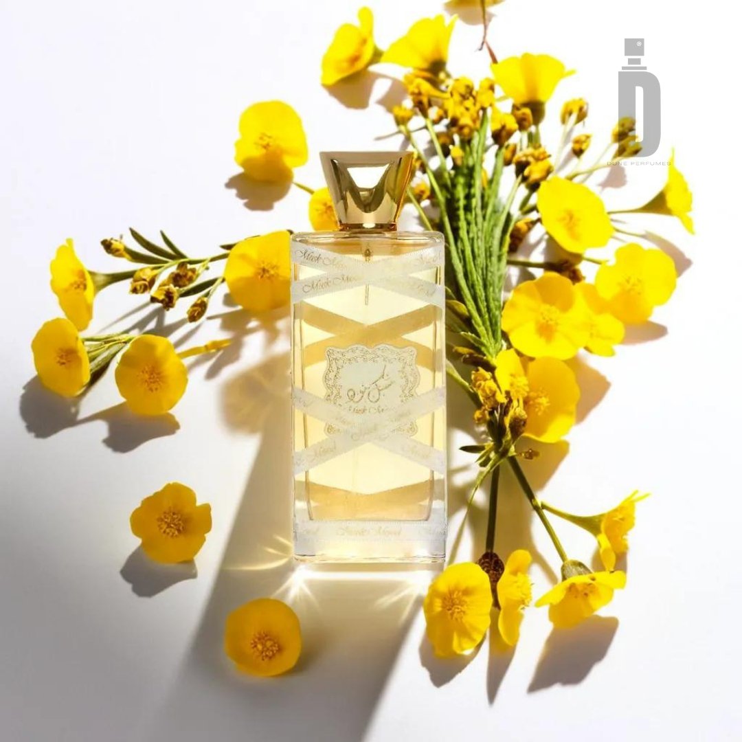 A bottle of Musk Mood 100ml EDP by Lattafa with a gold cap sits amidst a cluster of vibrant yellow flowers on a white background. The perfume liquid inside the clear bottle appears light yellow, emitting an aura of timeless allure. The scene is brightly lit, highlighting the floral decor and the fragrance's soft shadow.