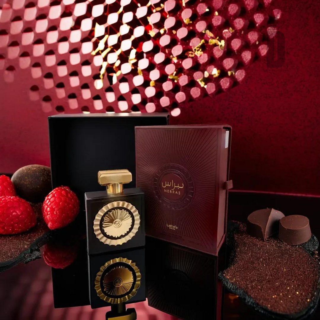 An elegant perfume bottle with a gold and black design, labeled "Nebras 100ml EDP - Lattafa Pride," sits beside a matching ornate box. The setup, showcasing the Nebras fragrance by Lattafa, is decorated with raspberries, chocolates, and a luxurious red textured background to ignite romance.