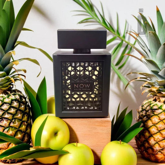 A black perfume bottle labeled "Now Rave 100ml EDP - Lattafa" from the brand Lattafa is surrounded by fresh green apples and whole pineapples. The bottle features an intricate, patterned design. In the backdrop, tropical leaves enhance the fresh and vibrant aesthetic of the image, promising an unforgettable olfactory journey.