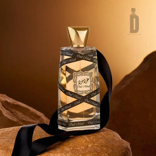 A bottle of Oud Mood 100ml EDP by Lattafa sits on a rock with a golden brown background. The luxurious depth of the perfume is highlighted by its golden cap and black ribbon featuring the text "Oud Mood," containing an amber-colored liquid. The Lattafa exotic fragrance logo is visible at the top right.