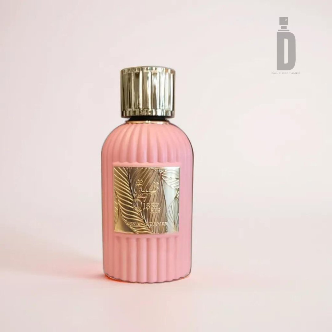 A pink, ribbed glass perfume bottle with a gold-colored metallic cap. The bottle, named Qissa Pink 100ml EDP by Paris Corner, features a decorative gold label with an ornate leaf design and some text. The background is a soft, pale pink, and the upper right corner has a subtle logo resembling a bottle that hints at its floral notes.