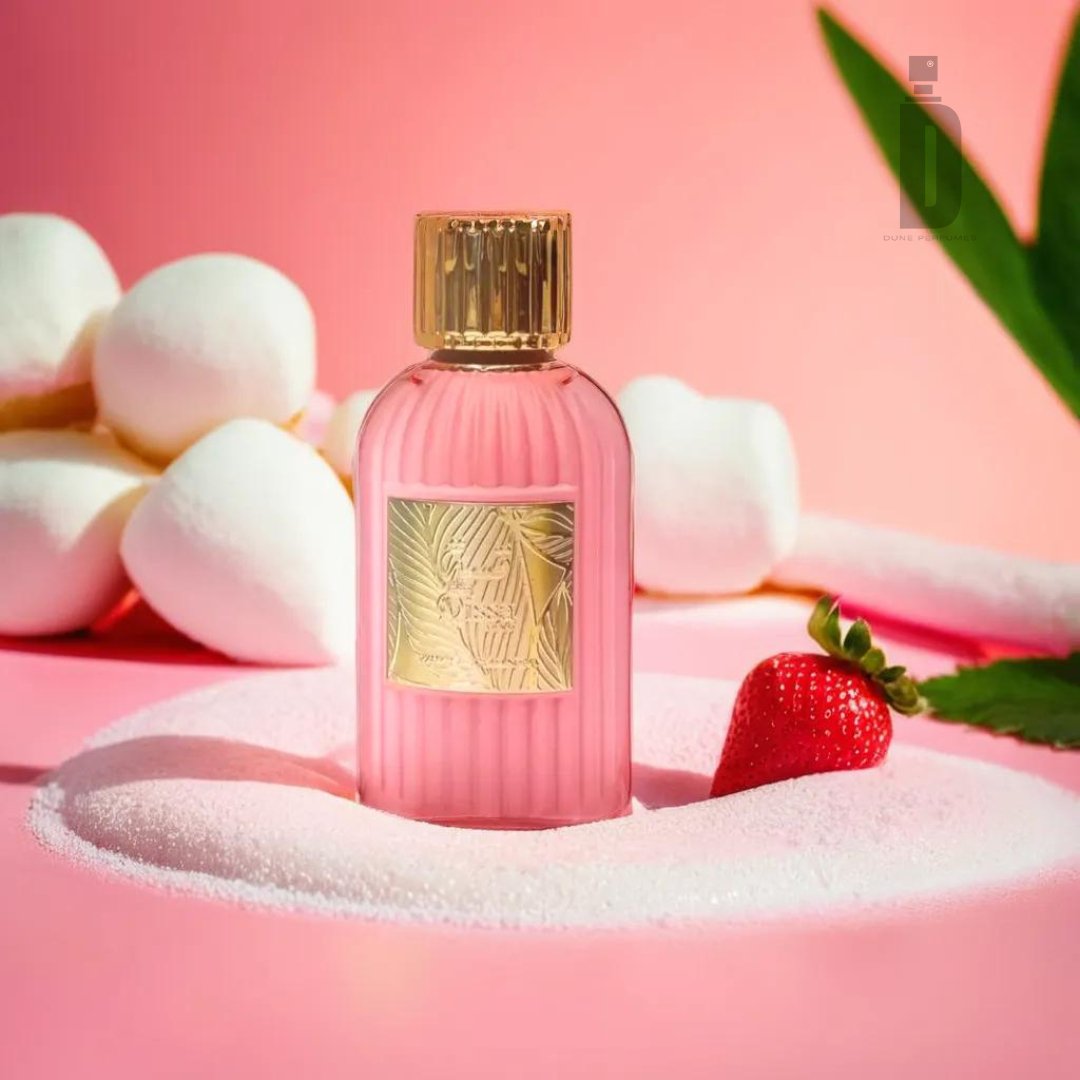 A pink Qissa Pink 100ml EDP bottle from Paris Corner, adorned with a gold cap and embossed with an intricate floral pattern, sits on a sugary surface alongside a whole strawberry. Marshmallows and green leaves are arranged in the background. The scene reflects the fragrance's floral notes against a playful pink backdrop, creating a sweet aesthetic.