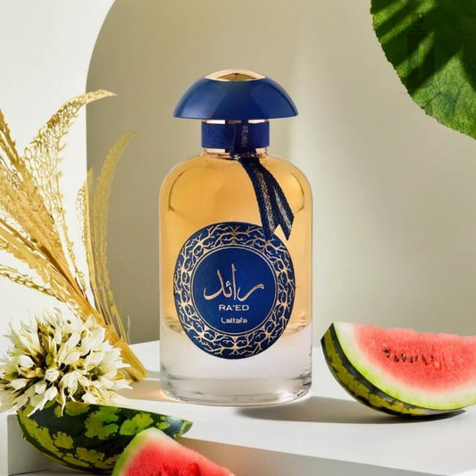 A 100ml bottle of "Ra'ed Luxe Gold" Eau de Parfum by Lattafa is showcased with an ornate blue cap and a coordinating blue and gold label. The luxurious bottle is surrounded by decorative foliage, a white flower, and sliced watermelon against a softly lit white and green backdrop.