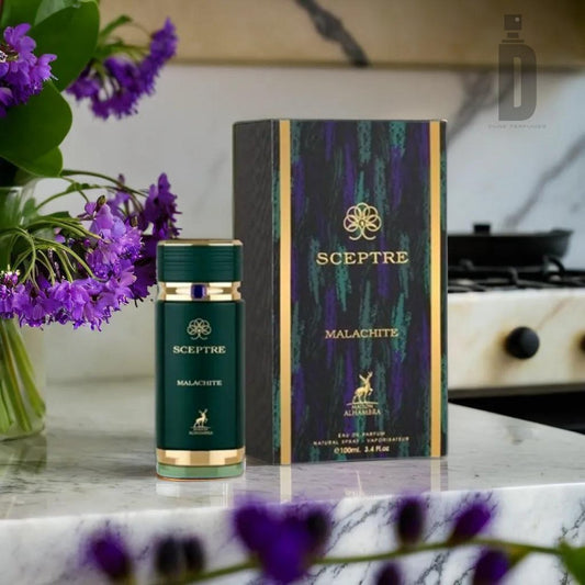 A bottle of Sceptre Malachite 100ml EDP from Maison Alhambra is placed on a kitchen counter next to its box. The green and gold accents of the bottle and box create a harmonious composition. In the background, there's a stove, and on the left, a vase with purple flowers adds a touch of elegance to the scene.