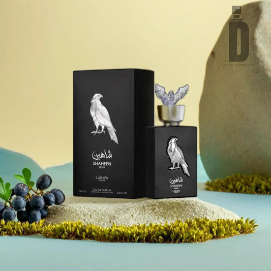 A bottle of Shaheen Silver 100ml Eau de Parfum by Lattafa Pride, adorned with Arabic text and a silver falcon graphic, rests on moss. Beside it is its matching black box showcasing the same falcon design. The elegant display includes a rock and some berries against a soft yellow-green backdrop, exuding pure sophistication.