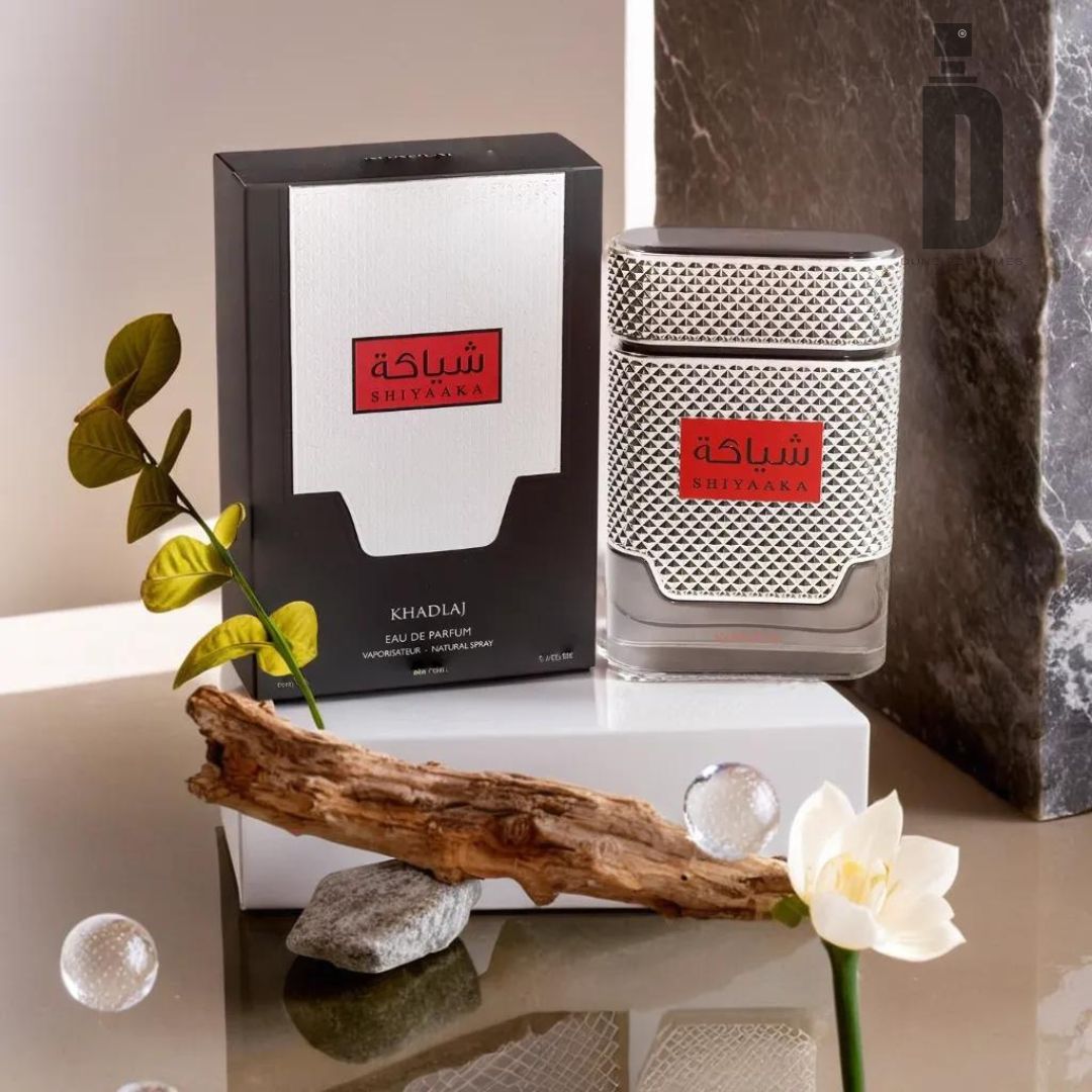 The Shiyaaka For Men 100ml EDP by Khadlaj is showcased in a stylish bottle, along with its box adorned with Arabic text. The display is complemented by natural decorative elements such as a small branch, a blossoming flower, and green leaves. The background features a blend of smooth and textured surfaces for a modern aesthetic.