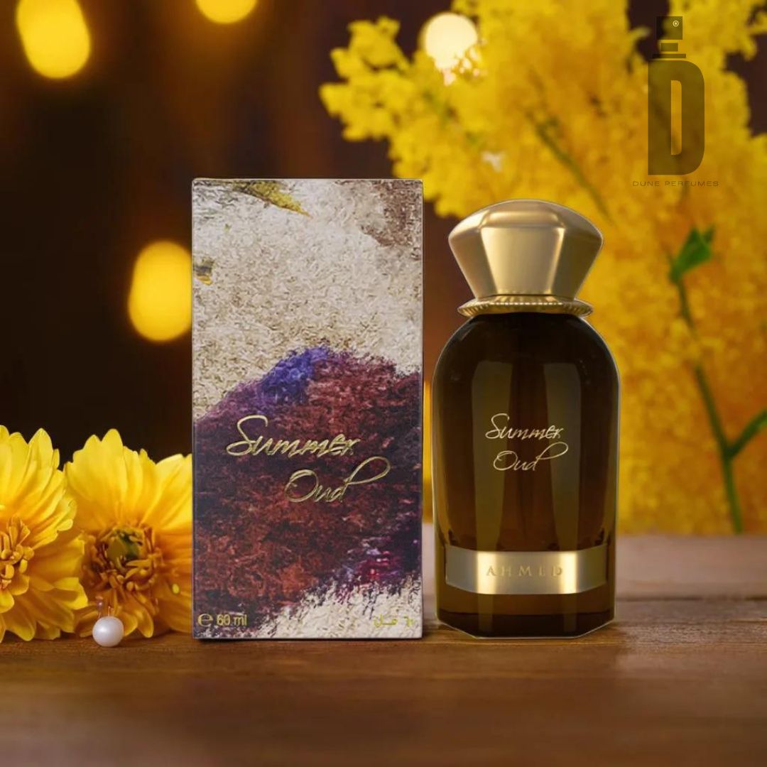 A bottle of "Summer Oud 60ml EDP - Ahmed Al Maghribi" is displayed next to its box on a wooden surface. The box features an abstract design with earthy tones, while yellow flowers in the background create a warm and inviting atmosphere that perfectly complements the fragrance notes by Ahmed Al Maghribi.