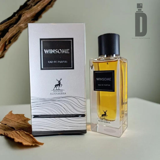 The image features a boxed and bottled perfume named "Winsome (The Tux)" 90ml Eau de Parfum by Maison Alhambra. The box and bottle are placed on a light surface with dry leaves and a wooden stick nearby. The sophisticated blend inside the transparent bottle, capped in black, is encased in an elegant white box with black accents.