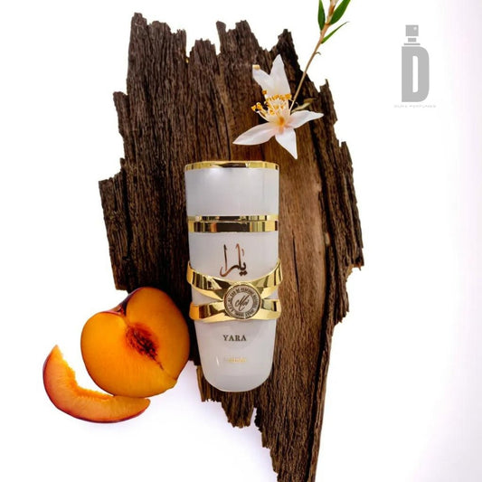 A luxurious presentation of the Yara Moi 100ml EDP by Lattafa is displayed on a piece of textured wood. The fragrance features a sleek white bottle with elegant gold accents, accompanied by a sliced peach and a delicate white flower. The brand's logo and name are prominently visible in grey at the top right corner.