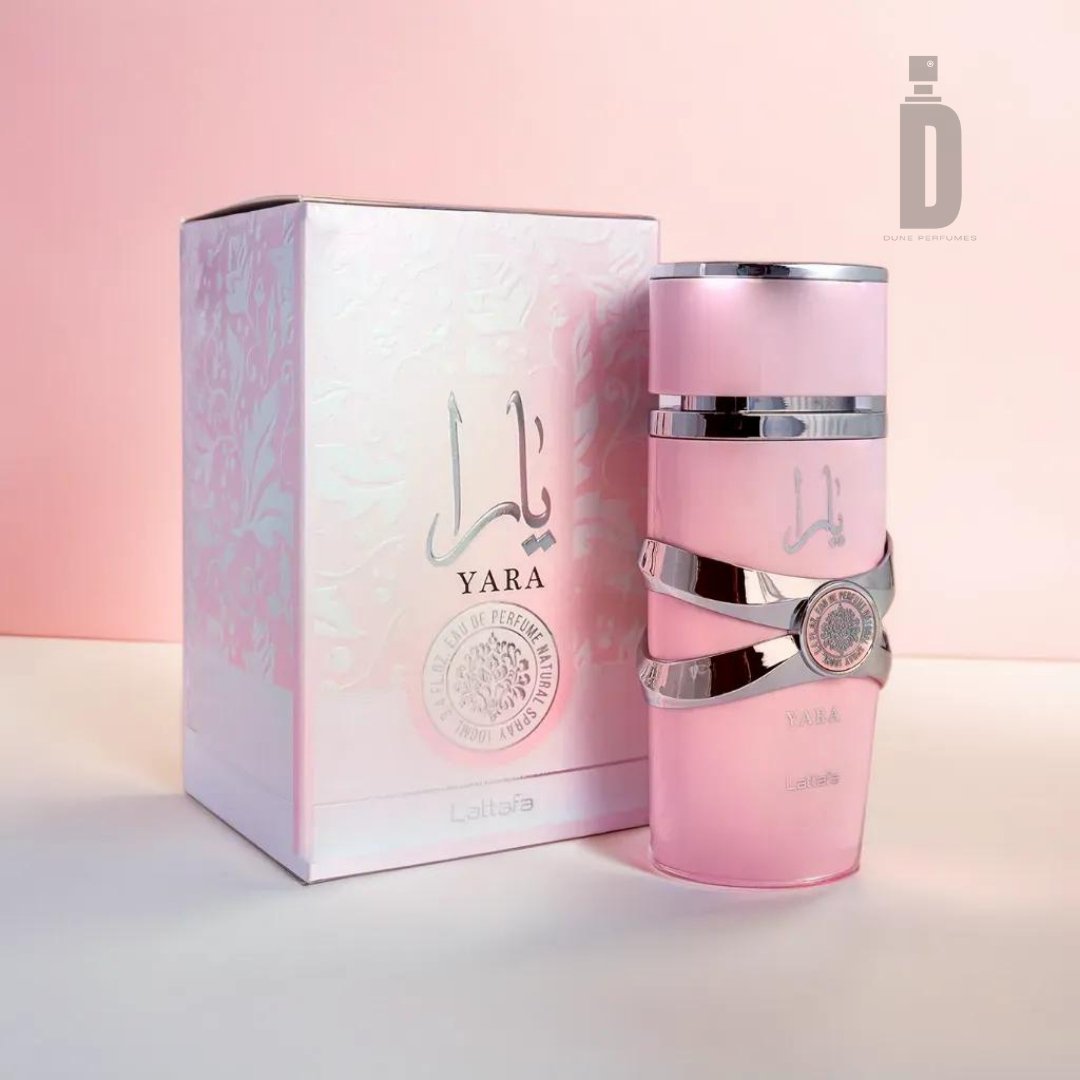 A Yara Pink 100ml EDP bottle by Lattafa, featuring silver accents and a pink hue, is positioned in front of its corresponding box that boasts an exquisite floral design interwoven with Arabic script. Embracing femininity, the Lattafa logo adorns both the box and the bottle. The soft gradient of pink tones in the background highlights its elegance.