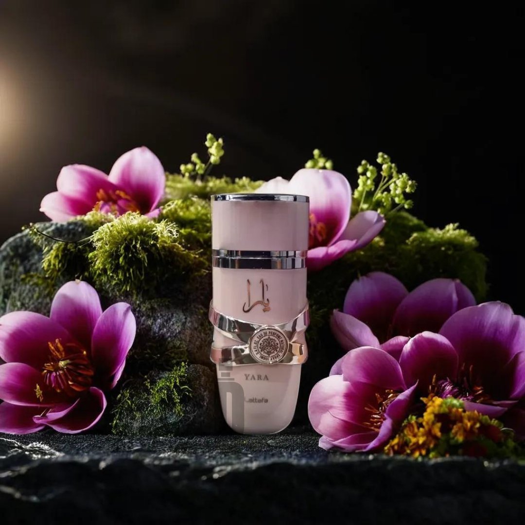 A sleek bottle of Yara Pink 100ml EDP by Lattafa is centered on a mossy rock surrounded by vibrant fuchsia and purple flowers, set against a dark, blurred background. The bottle features elegant silver accents and fine detail enhancing its luxurious appearance, radiating femininity and allure.