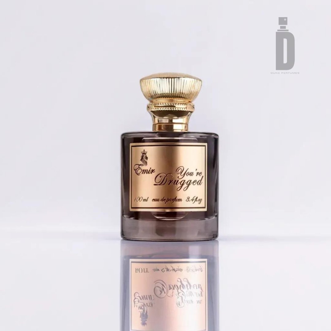 A perfume bottle labeled "You're Drugged 100ml EDP - Emir" from Paris Corner, with a dark, rounded body and a gold cap, sits against a plain white background. The bottle's reflection is visible on the surface below it, embodying its mesmerizing fragrance. A logo with the letter "D" is in the top right corner.